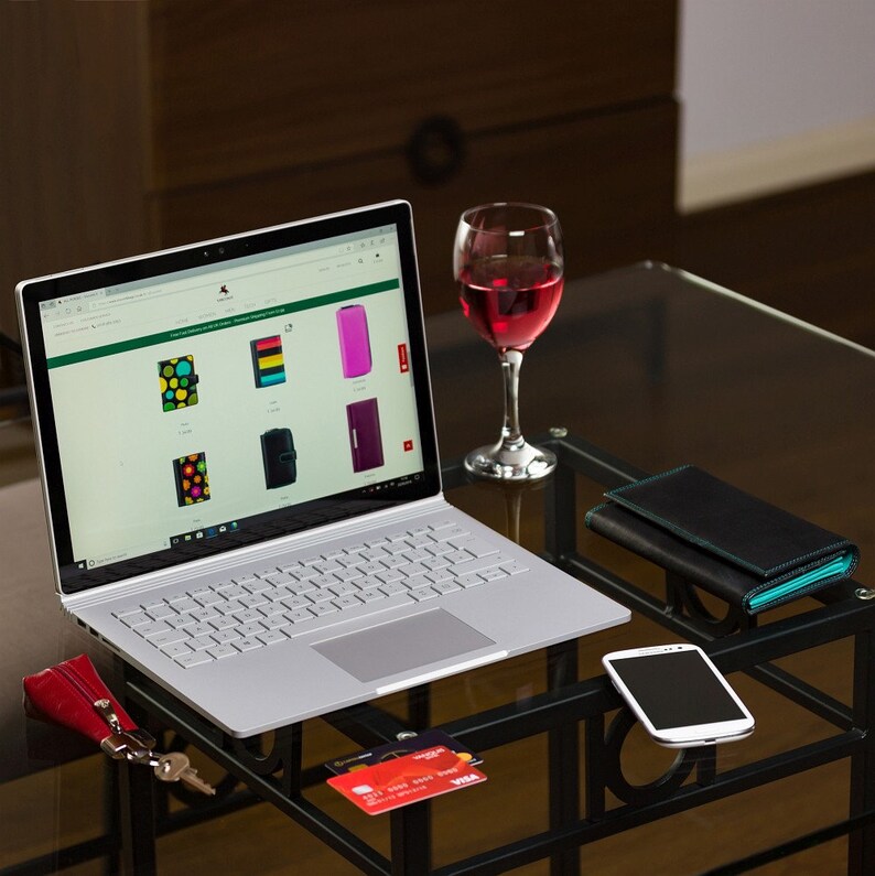 Pu&ograve; includere: Un computer portatile argentato con una tastiera bianca &egrave; aperto su un sito web che mostra custodie per telefono colorate. Sul tavolo ci sono anche un bicchiere di vino rosso, un portafoglio nero, una carta di credito e uno smartphone.
