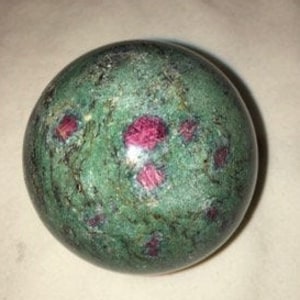 May include: A polished sphere of green zoisite with visible ruby inclusions. The sphere has a mottled appearance with varying shades of green and small, vibrant red ruby crystals scattered across its surface. The gemstone is smooth and rounded.