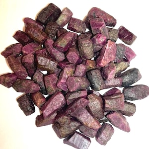 May include: A collection of raw, unpolished ruby gemstones in various shades of deep magenta, dark red, and purplish-pink. These natural mineral crystals feature irregular, jagged shapes with rough, textured surfaces and opaque to translucent finishes.