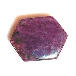 May include: A hexagonal, polished gemstone with a deep magenta hue. The stone has subtle variations in color and texture, with darker inclusions and lighter streaks. The edges are well-defined, and the overall appearance is smooth and reflective.