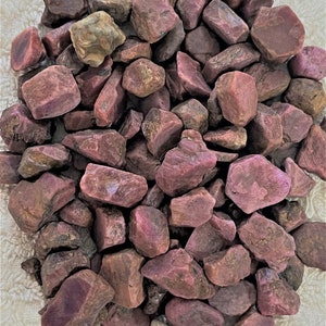 May include: A pile of rough, uncut ruby gemstones. The stones vary in size and shape, displaying a range of reddish-purple hues. Some stones have a slightly lighter, brownish tone. The gemstones are scattered on a white, textured surface.