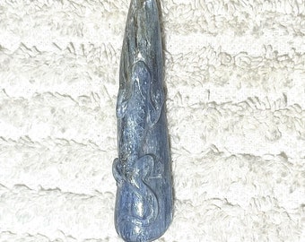 May include: A blue, elongated crystal pendant with carved details. The pendant has a smooth, polished surface and features a design of stylized figures. The crystal's color ranges from light to dark blue, with some areas appearing almost gray.