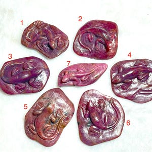 May include: Seven carved ruby lizard figurines. Each figurine is a different shape and size, with a detailed lizard carved into the surface. The ruby has a deep red color with some variations in tone. The lizards are in various poses.