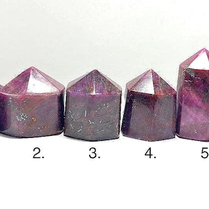 May include: Six polished ruby crystal towers with faceted tops. The crystals are a deep reddish-purple color, with variations in tone and visible natural inclusions. Each tower is numbered from one to six.