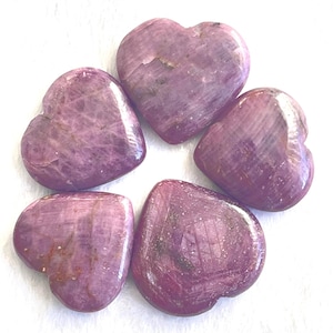 May include: Five heart-shaped stones in varying sizes, arranged on a white surface. The stones are a deep pinkish-purple color with natural variations in tone and texture. The polished surfaces catch the light, highlighting their smooth, rounded forms.