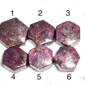 May include: Six hexagonal ruby gemstones in shades of pink and red, numbered 1 to 6. The polished stones display inclusions and dark veins. The gemstones are arranged on a white surface. Each stone is approximately 2-3 cm across.