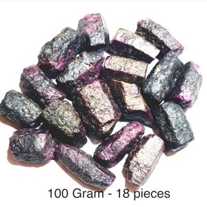 May include: A collection of 18 raw, dark purple and metallic grey ruby crystal stones with a rough, textured surface. The stones are grouped together, and the text 100 Gram - 18 pieces is printed clearly at the bottom of the image.