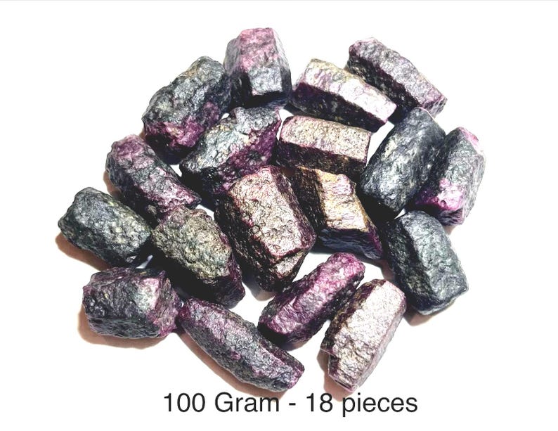 May include: A collection of 18 raw, dark purple and metallic grey ruby crystal stones with a rough, textured surface. The stones are grouped together, and the text 100 Gram - 18 pieces is printed clearly at the bottom of the image.