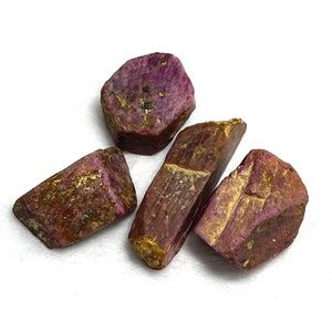 May include: Four rough ruby gemstones with a deep pinkish-red hue and golden-brown accents. The raw, uncut stones have irregular shapes and varying sizes, showcasing natural textures and crystalline structures. The gemstones are set against a white background.