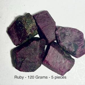 May include: Five raw ruby gemstones with deep magenta and dark red hues, featuring natural crystalline textures and irregular geometric shapes. The collection of stones has a combined weight of 120 grams as indicated by the text Ruby - 120 Grams - 5 pieces.
