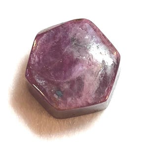 May include: A faceted, hexagonal gemstone with a deep magenta hue. The polished surface reflects light, revealing internal patterns and variations in color. The gem is set against a white background, highlighting its rich color and form.