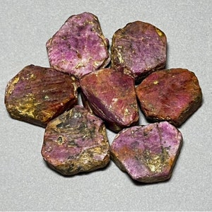 May include: A collection of seven rough-cut ruby gemstones. The stones display a deep red-purple hue with areas of gold and brown. Each stone has a unique, irregular shape, showcasing natural textures and variations.