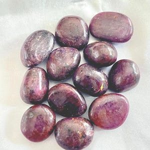 May include: A collection of polished, oval-shaped gemstones in varying sizes. The stones display a deep burgundy color with subtle variations in tone and texture. They are arranged on a white, slightly textured surface.
