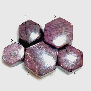 May include: Five hexagonal, polished ruby gemstones are arranged on a light background. The rubies vary in size and have a deep red-purple hue. Each stone has a unique pattern of internal lines and inclusions, with a reflective surface.