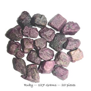 May include: A collection of twenty raw ruby stones with deep magenta and dark purple hues. The stones have irregular, faceted shapes and varied textures. Text at the bottom reads: Ruby - 127 Grams - 20 pieces. These natural gemstones are displayed together.