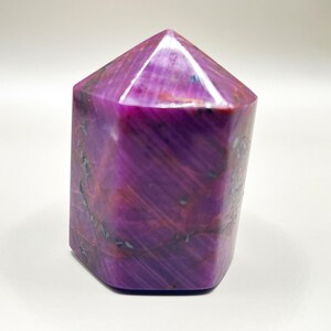 Puede incluir: Una gema pulida y facetada en tonos de morado intenso y magenta. El cristal tiene una base rectangular y una parte superior piramidal. La superficie de la piedra muestra vetas naturales y variaciones de color, creando una apariencia única. La gema está sobre un fondo blanco.