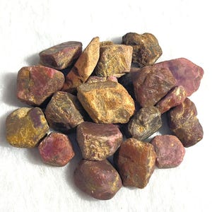 May include: A collection of raw, unpolished ruby gemstones in various sizes and shapes. The stones exhibit a mix of deep red, maroon, and earthy brown tones with natural, rough textures and irregular, angular facets across their surfaces.