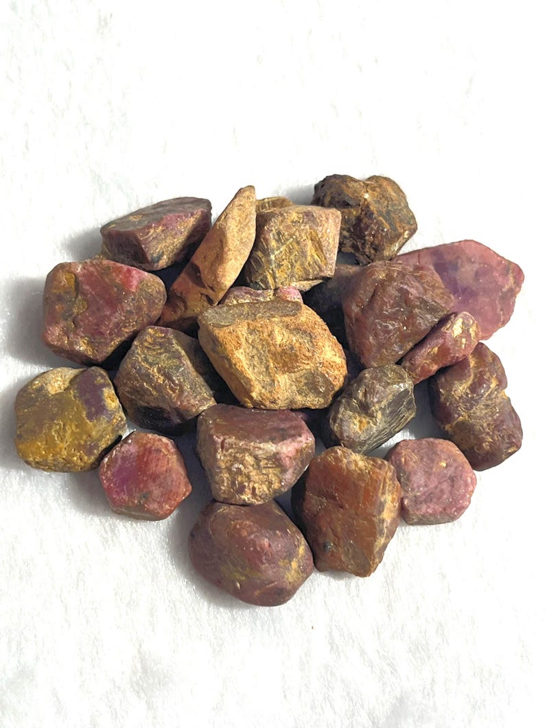 May include: A collection of raw, unpolished ruby gemstones in various sizes and shapes. The stones exhibit a mix of deep red, maroon, and earthy brown tones with natural, rough textures and irregular, angular facets across their surfaces.