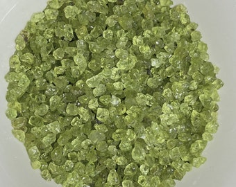 May include: A close-up view of a white bowl filled with small, irregularly shaped, light green peridot gemstones. The gemstones are translucent and reflect light, creating a sparkling effect. The bowl is white and provides a neutral background for the gemstones.