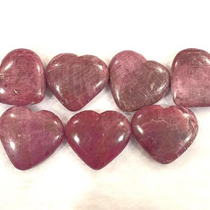 May include: Seven polished heart-shaped stones in varying shades of pink and red. The stones have a smooth, glossy finish and natural veining patterns. The hearts are arranged on a white surface.