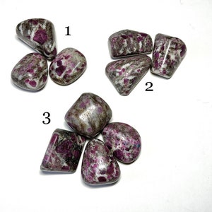 May include: A collection of tumbled stones with a mix of gray, white, and deep pink hues. The stones have a speckled appearance, with the pink color concentrated in various spots. The stones are smooth and rounded, with a natural, earthy aesthetic.