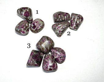 May include: A collection of tumbled stones with a mix of gray, white, and deep pink hues. The stones have a speckled appearance, with the pink color concentrated in various spots. The stones are smooth and rounded, with a natural, earthy aesthetic.
