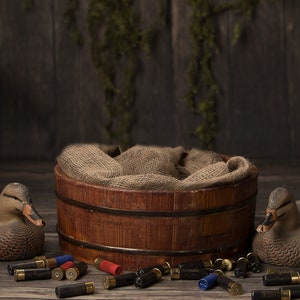 Op de afbeelding: Een rustieke houten ton met jute voering, geflankeerd door twee eendenlokvogels en verspreide hagelpatronen. De scène speelt zich af op een verweerd houten oppervlak, met een donkere achtergrond en groen.