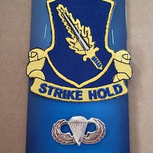 May include: A blue and yellow embroidered patch with the words "STRIKE HOLD" and a sword and leaf design. Below the patch is a silver parachute and wing pin. The top of the card reads "BIGUNS PATCHES & PINS & MORE."