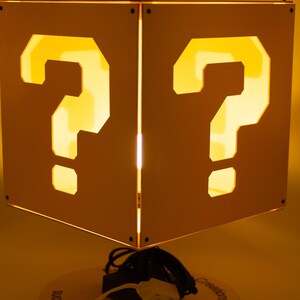 May include: A square lamp with white panels featuring cut-out question marks that glow with a warm yellow light. The lamp is mounted on a circular base with the text Beyond Basic Creations printed on it, and includes a white power cord and plug.