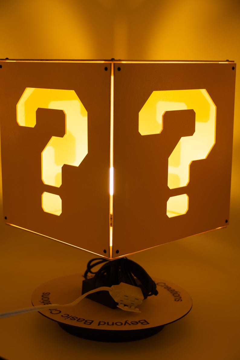 May include: A square lamp with white panels featuring cut-out question marks that glow with a warm yellow light. The lamp is mounted on a circular base with the text Beyond Basic Creations printed on it, and includes a white power cord and plug.