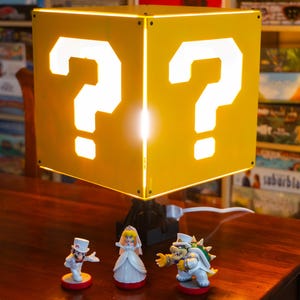 May include: A yellow cube-shaped lamp featuring a white question mark cutout on each visible side. The lamp is illuminated from within and sits on a black base. Three small character figurines are positioned in front of the lamp base.