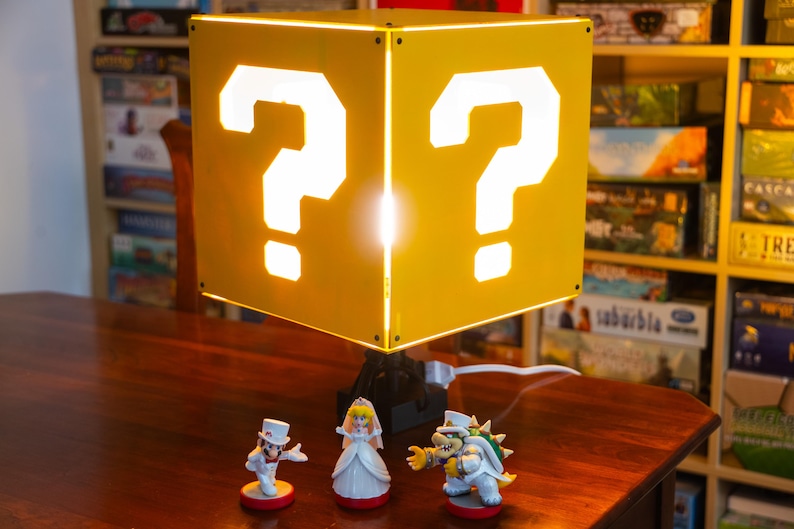 May include: A yellow cube-shaped lamp featuring a white question mark cutout on each visible side. The lamp is illuminated from within and sits on a black base. Three small character figurines are positioned in front of the lamp base.