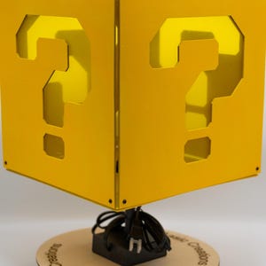 May include: A yellow cube-shaped lamp featuring a cutout question mark on each visible side. The lamp is mounted on a circular wooden base with the text "Beyond Basic Creations" printed on it, and includes a black power cord and plug.