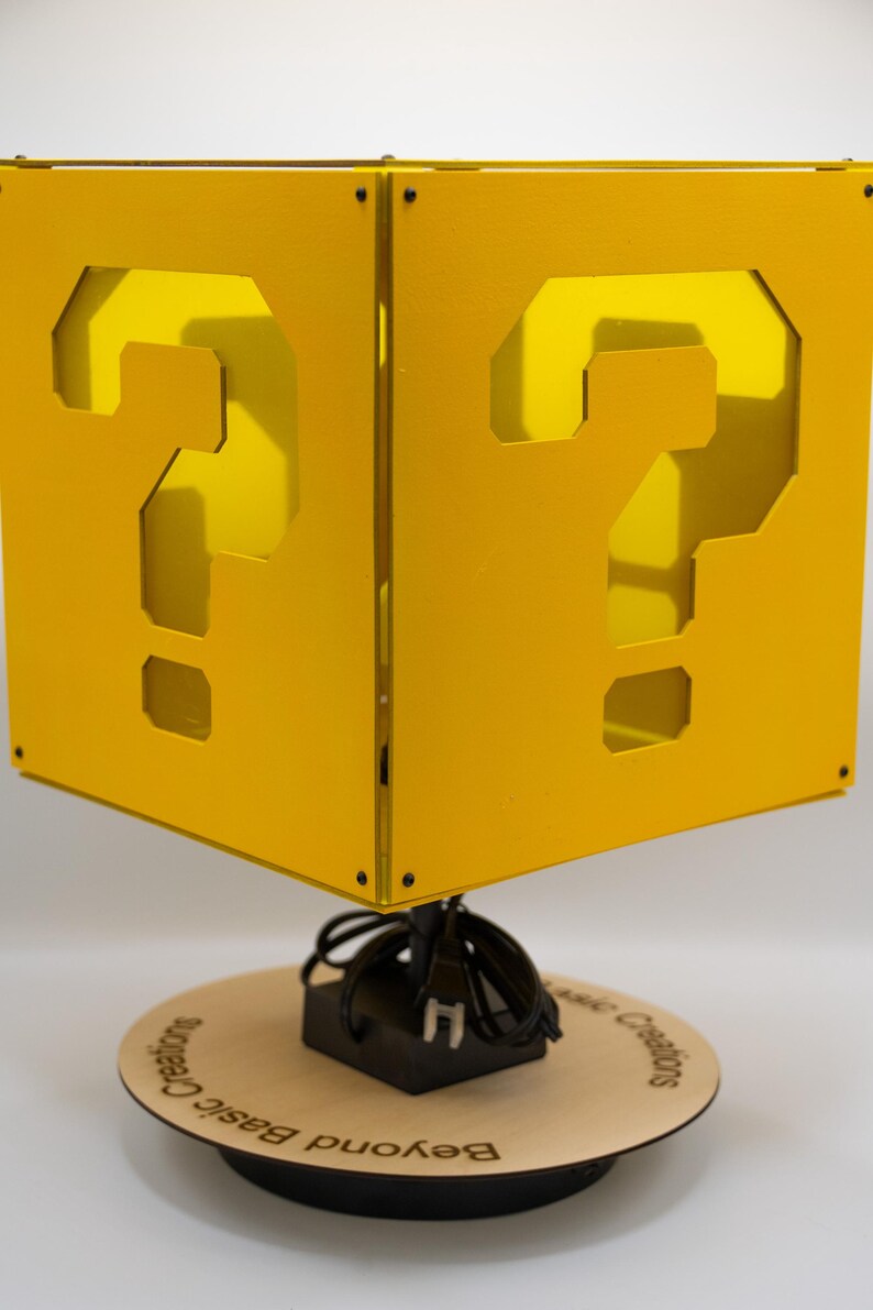 May include: A yellow cube-shaped lamp featuring a cutout question mark on each visible side. The lamp is mounted on a circular wooden base with the text "Beyond Basic Creations" printed on it, and includes a black power cord and plug.