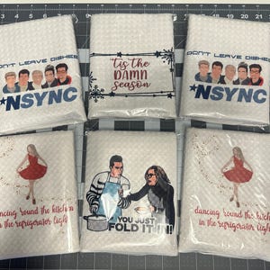 May include: Six white kitchen towels with different designs. The towels feature a band with the text "Don't Leave Dishes" and "*NSYNC", a Christmas-themed design with the text "Tis the Damn Season", a design with a couple folding a dish towel with the text "You Just Fold It In!" and two designs with a woman dancing with the text "Dancing Round the Kitchen in the Refrigerator Light".