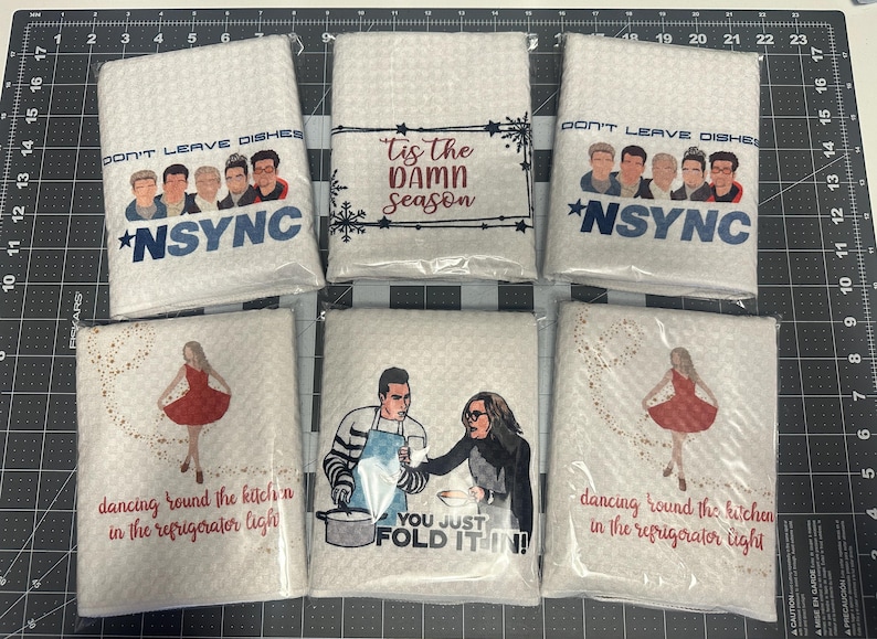 May include: Six white kitchen towels with different designs. The towels feature a band with the text "Don't Leave Dishes" and "*NSYNC", a Christmas-themed design with the text "Tis the Damn Season", a design with a couple folding a dish towel with the text "You Just Fold It In!" and two designs with a woman dancing with the text "Dancing Round the Kitchen in the Refrigerator Light".