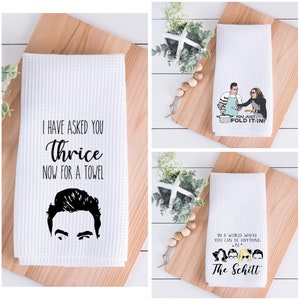 Puede incluir: Tres toallas de cocina blancas con diferentes diseños. La primera toalla presenta una ilustración en blanco y negro de la cara de un hombre con el texto "Te he pedido tres veces una toalla". La segunda toalla presenta una ilustración de dibujos animados de dos personas con el texto "¡Solo dóblala!". La tercera toalla presenta una ilustración de dibujos animados de un grupo de personas con el texto "En un mundo donde puedes ser cualquier cosa, sé The Schitt".