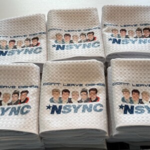 May include: Six white kitchen towels with a blue and white design. The towels feature a group of five cartoon men with the text "Don't Leave Dishes *NSYNC" printed on them.