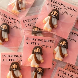 May include: A collection of small Jesus figurines, each individually packaged on pink cards. The cards feature the text "EVERYONE NEEDS A LITTLE Jesus" in black script. The figurines are white, brown, and tan.