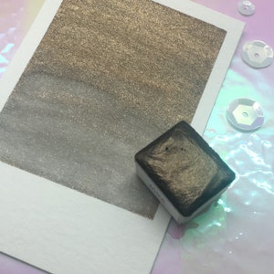 以下が含まれることがあります： 金色と銀色の水彩で描かれた白いカード。カードの右下隅には、小さな黒い金色の水彩絵の具の容器があります。容器には「KWA」と印刷されています。