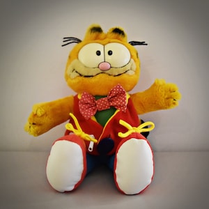 Puede incluir: Un juguete de peluche naranja Garfield que lleva un chaleco rojo con una pajarita amarilla y zapatos blancos. El juguete tiene una nariz negra y bigotes.