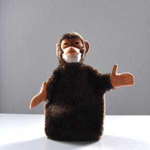 May include: A brown plush monkey puppet with a light brown face, ears, and hands. The puppet has a white beard and is posed with its arms outstretched, thumbs up. The monkey puppet is set against a gray background.