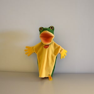 Puede incluir: Marioneta de mano de rana verde y amarilla con una gran boca abierta. La marioneta lleva una camisa amarilla con una etiqueta roja que dice "Original Hand Puppets Made in Germany".