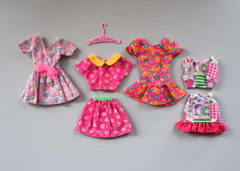 May include: A set of six doll clothes in pink, green, yellow, and white with floral and geometric patterns. The clothes include a dress, a skirt, a top, a dress, a top, and a skirt.