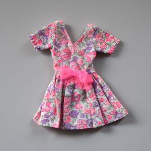 May include: A pink and purple floral print dress with short sleeves and a pink bow at the waist. The dress is made for a doll.