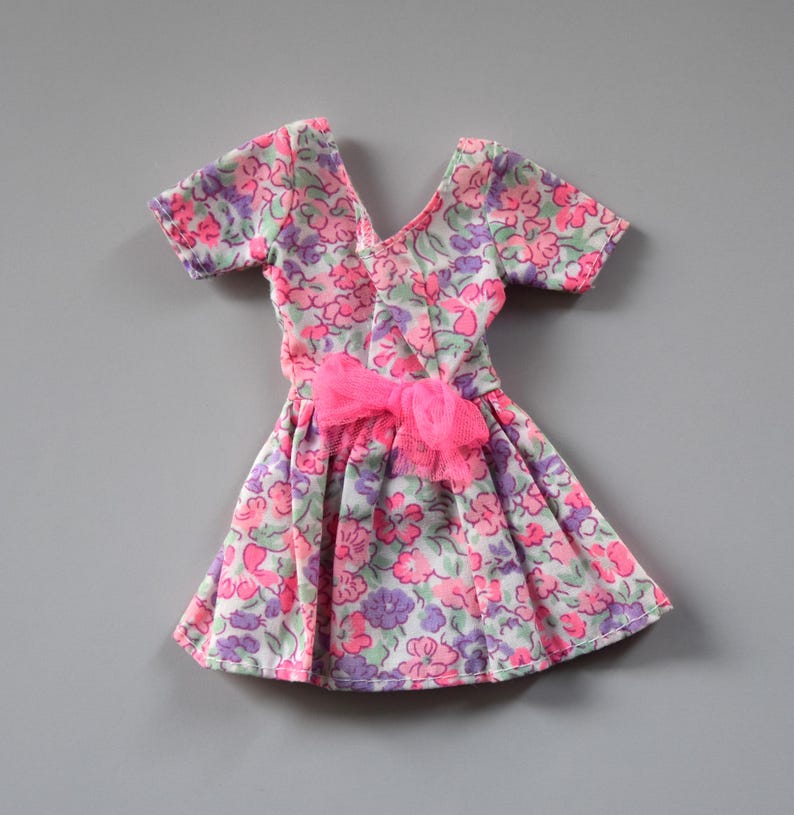 May include: A pink and purple floral print dress with short sleeves and a pink bow at the waist. The dress is made for a doll.