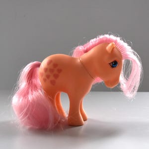 Puede incluir: Un poni de juguete vintage de color melocotón con crin y cola rosas. El poni tiene ojos azules y marcas en forma de corazón en el flanco. Un artículo de colección.