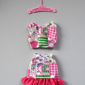 May include: A pink plastic hanger with a doll-sized two-piece outfit. The outfit is made of a patchwork fabric with pink, green, and white squares and a floral pattern. The skirt has a pink ruffle.