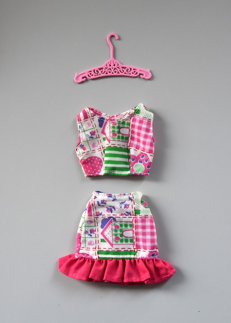 May include: A pink plastic hanger with a doll-sized two-piece outfit. The outfit is made of a patchwork fabric with pink, green, and white squares and a floral pattern. The skirt has a pink ruffle.