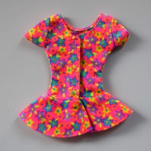 May include: A pink, yellow, blue, and green doll dress with a star print. The dress has short sleeves and a gathered skirt.